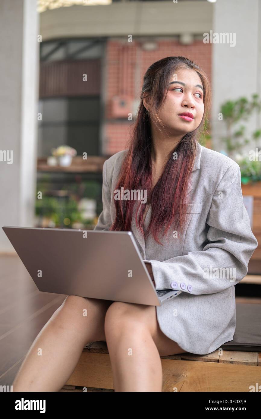 Eine vertikale Mittelaufnahme einer jungen südostasiatischen thailändischen Isan-Geschäftsfrau, die im Freien sitzt, mit einem silbernen Laptop aufgeschlagen auf ihrem Schoß, und die weg von ihr schaut Stockfoto