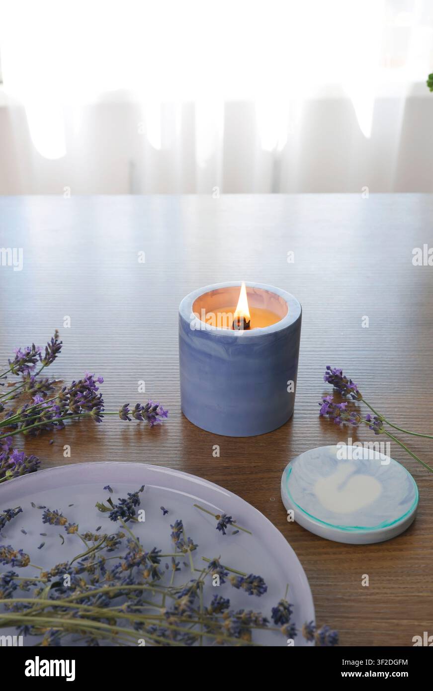 Duftende Soja Kerze brennt in blauem Marmor-Betthalter auf Holz Esstisch in der Nähe eines Keramiktabletts mit getrockneten Lavendelblüten für einen gemütlichen Morgen Stockfoto