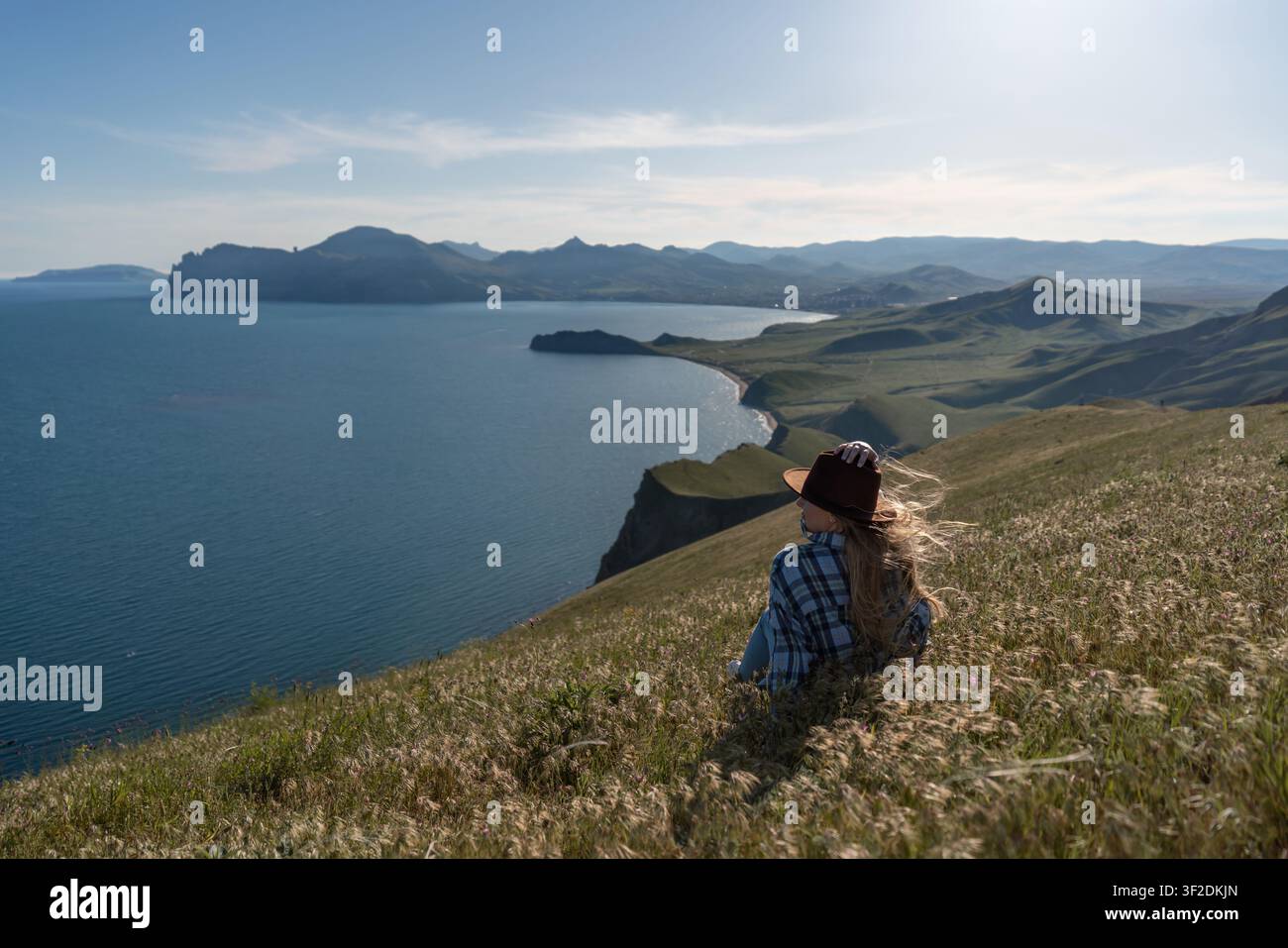 Frau hat Landschaft, Frau sitzt auf grasbewachsenem Hügel mit Blick auf Bucht und Berge an einem sonnigen Tag und genießt das friedliche Reiseziel Stockfoto