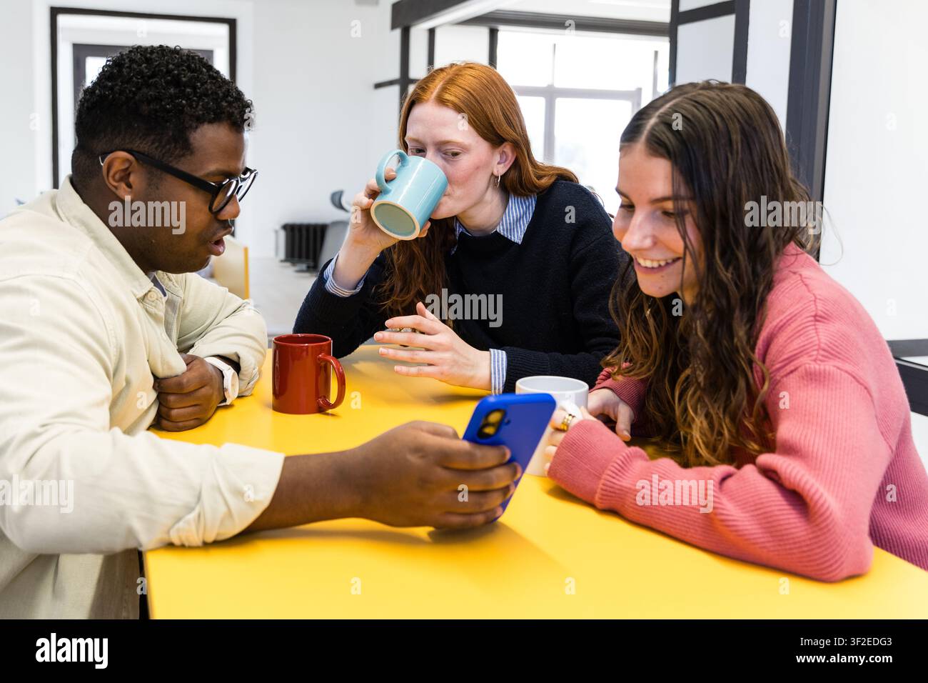 Drei Kollegen versammeln sich um einen gelben Tisch in einem modernen Büro, lächeln, während sie ein Smartphone betrachten und Kaffee aus Tassen schlürfen, während sie einen entspannten Tee genießen Stockfoto