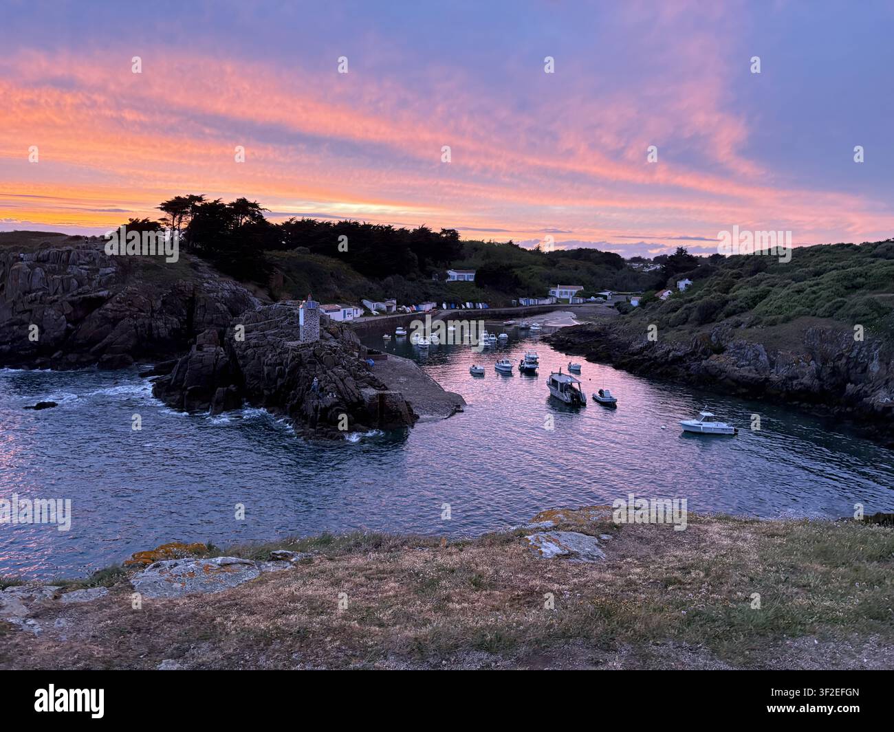 Sonnenuntergang über dem Hafen von Port de la Meule, Ile d'Yeu, Vendée, Frankreich. Fischerboote in einer felsigen Bucht, Steinturm-Leuchtturm, lebhafter rosa Himmel. - Smartphone-aufgenommenes Stockfoto