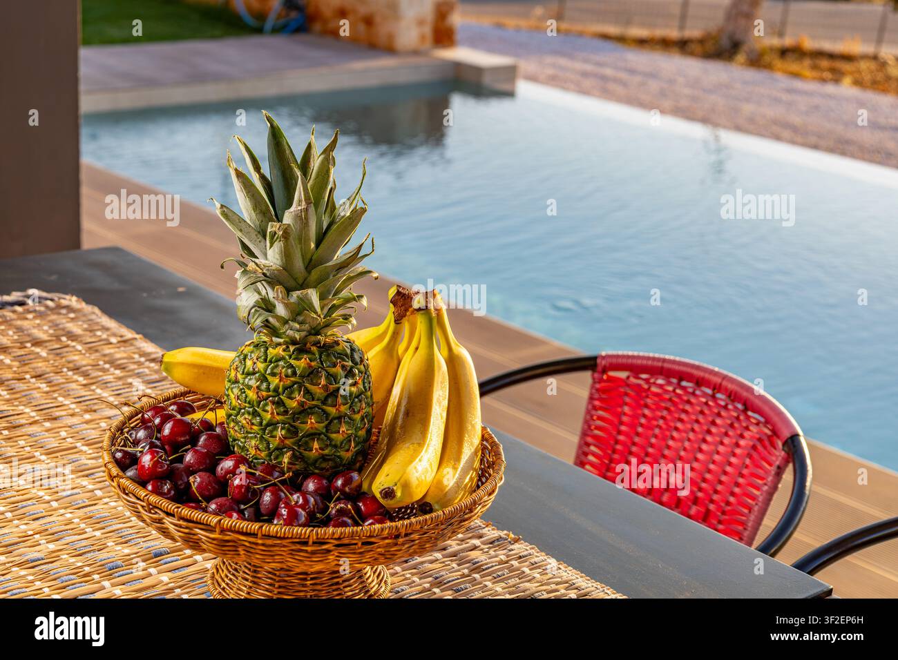 Ansicht eines Korbs gefüllt mit einer frischen Ananas, reifen Bananen und dunkelroten Kirschen auf einem Tisch neben einem Schwimmbad in Chania, Griechenland. Stockfoto