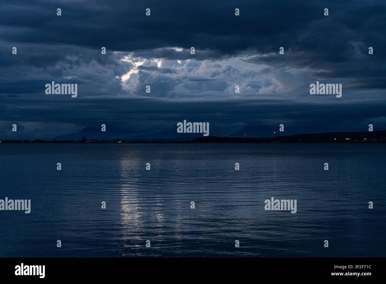 Durchbruch des Mondlichts durch dunkle Gewitterwolken, die sich auf ruhigem Wasser in der Dämmerung spiegeln, mit fernen Bergen und Uferlichtern Stockfoto