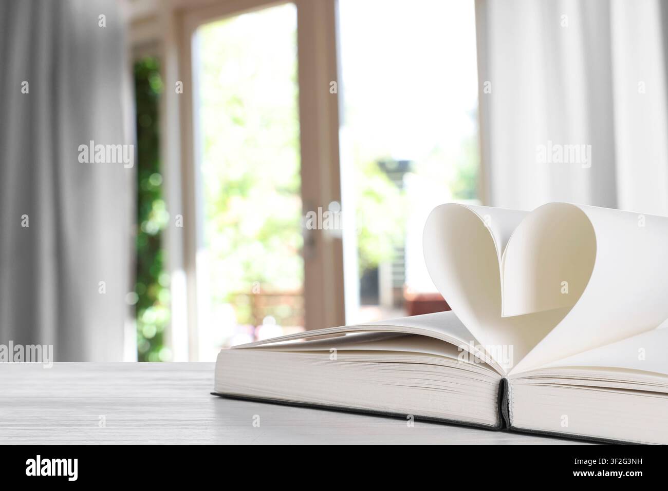 Buch mit Herz aus Seiten auf dem Tisch vor einem verschwommenen Fenster, Nahaufnahme. Platz für Text Stockfoto