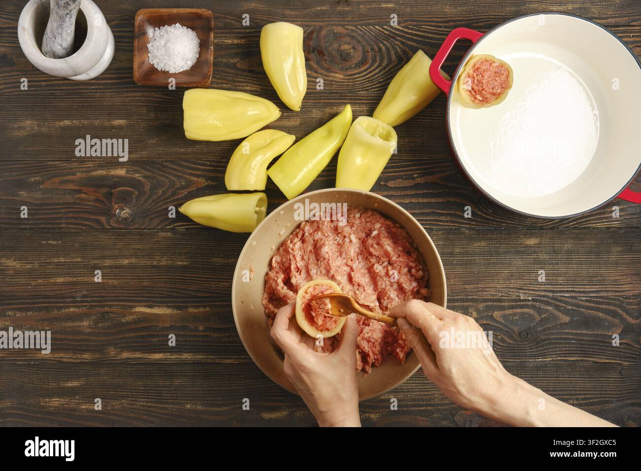 Hände füllen Paprika mit Hackfleisch. In der Nähe liegen halbierte Paprika, die gefüllt werden sollen. Der Küchentisch zeigt etwas Salz und einen gusseisernen Topf. Szene Stockfoto
