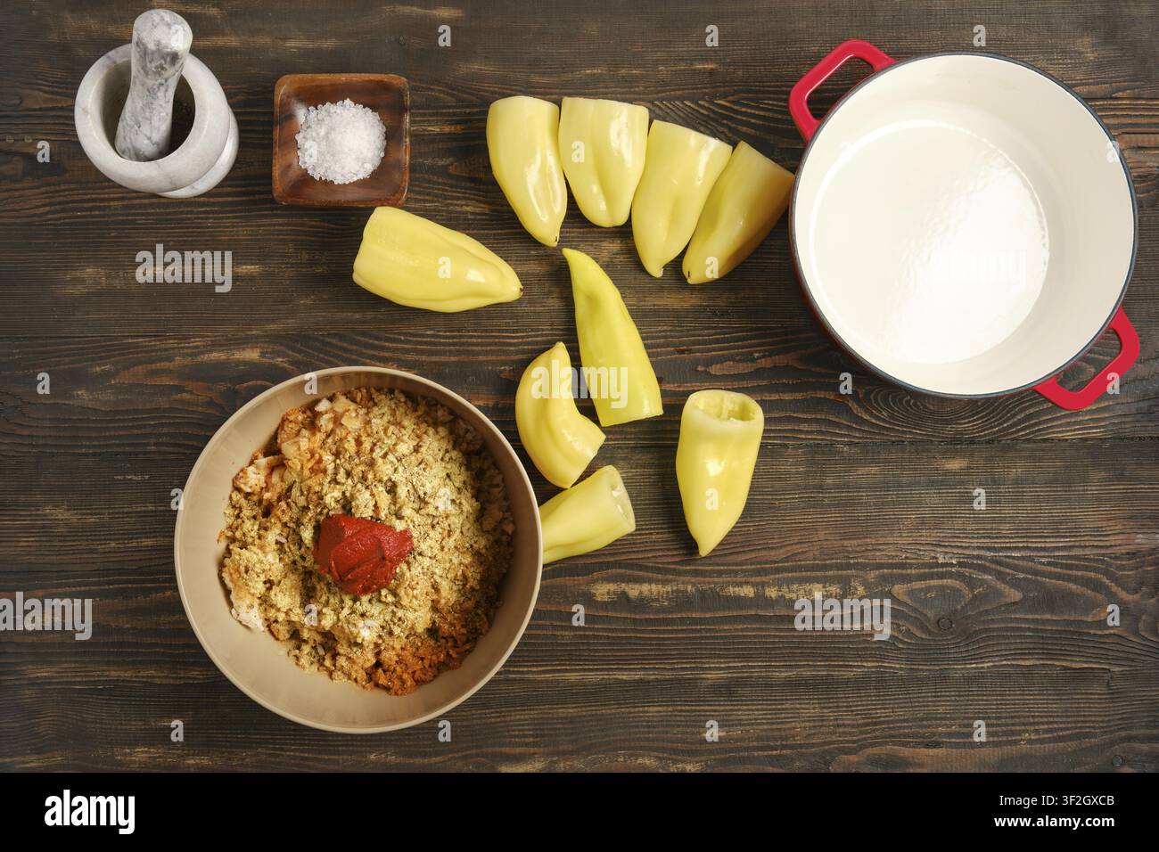 Die Zubereitung von gefüllten Paprika findet auf einem Holztisch statt. Die Zutaten umfassen gehackte Paprika, eine Schüssel mit Getreidemischung, Salz und einen Topf mit Milch. Ko Stockfoto