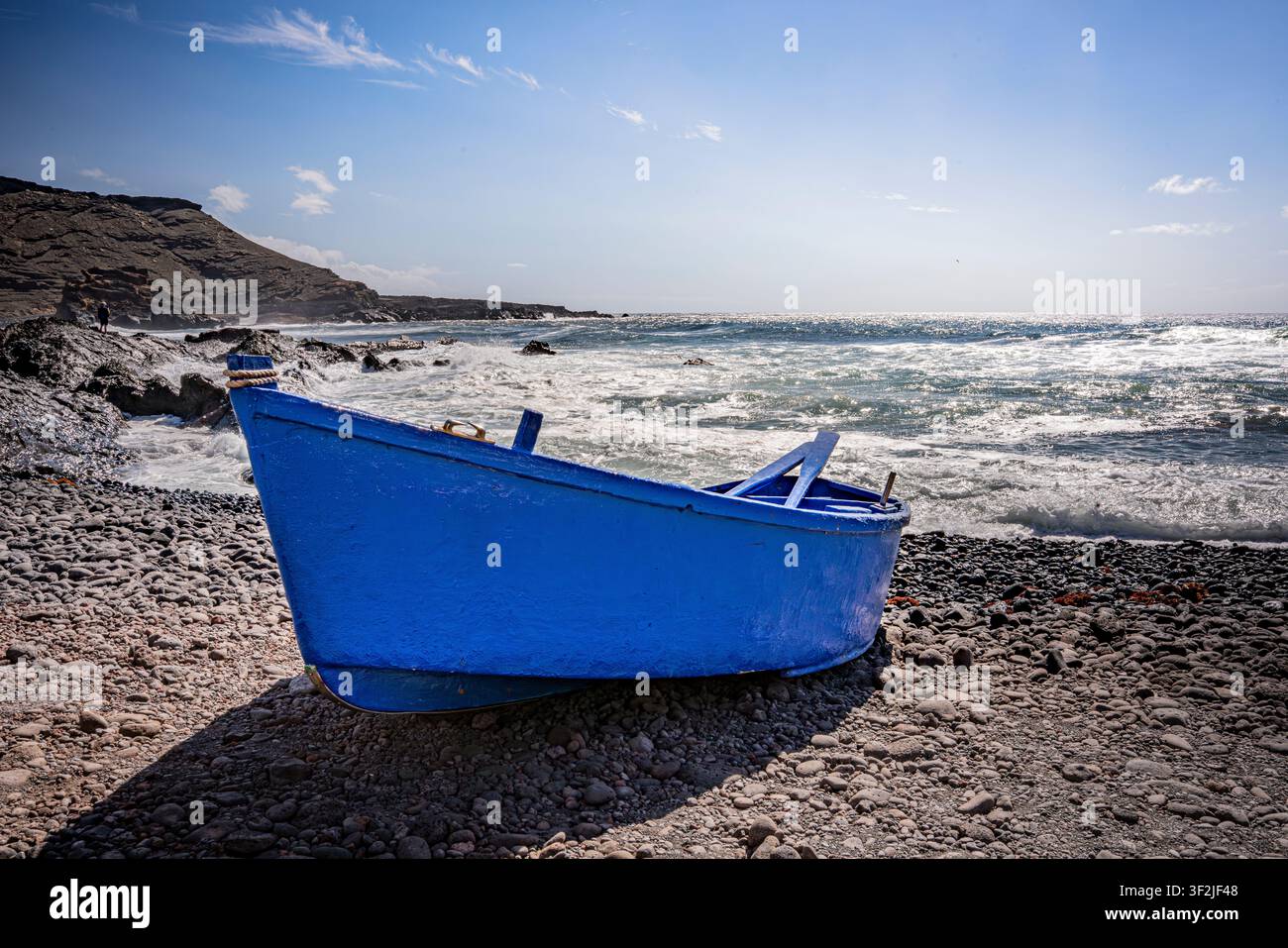 Blaues Boot ruht am Ufer auf einem vulkanischen Kiesstrand, während Ozeanwellen an die Küste schlagen Stockfoto