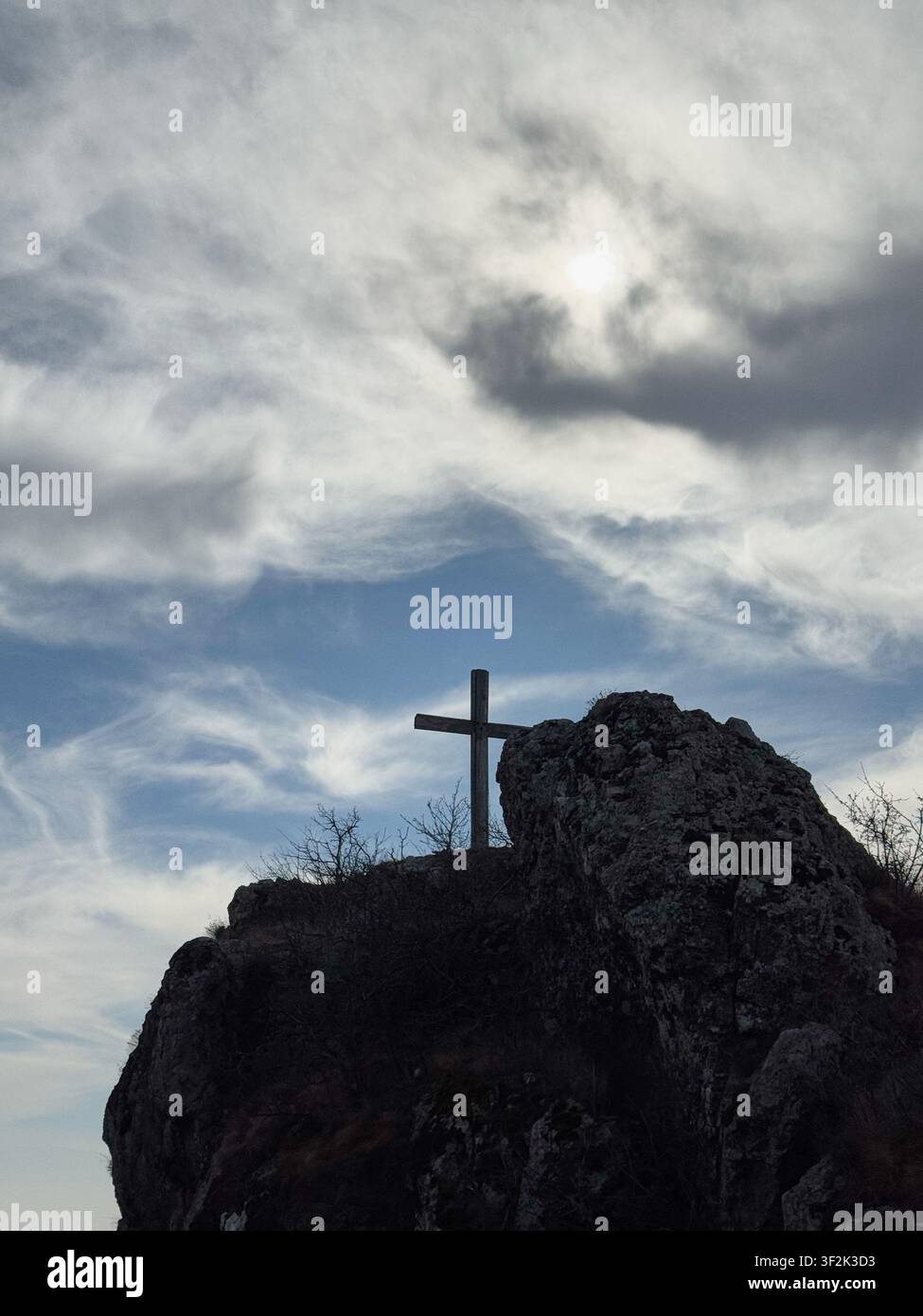 Kreuz auf felsigem Gipfel unter wolkigem Himmel mit hellem Sonnenlicht, das von den Wolken diffundiert. Stockfoto