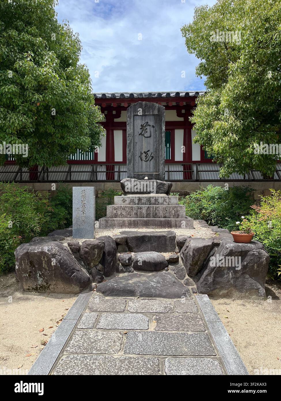 19. Juli 2023, Osaka, Japan – Stele aus Stein mit japanischen Kanji-Inschriften im Tempelhof, rote traditionelle Architektur und üppige Bäume im Hintergrund. - Smartphone-aufgenommenes Stockfoto