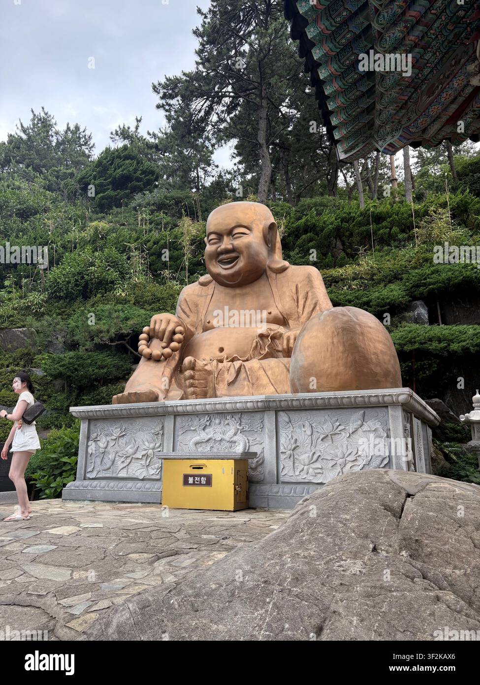 15. Juli 2025, Busan, Südkorea – Große Maitreya-lachende-Buddha-Steinstatue in einem buddhistischen Tempelhof mit traditionellem Pagoden-Dach und Kiefern. - Smartphone-aufgenommenes Stockfoto