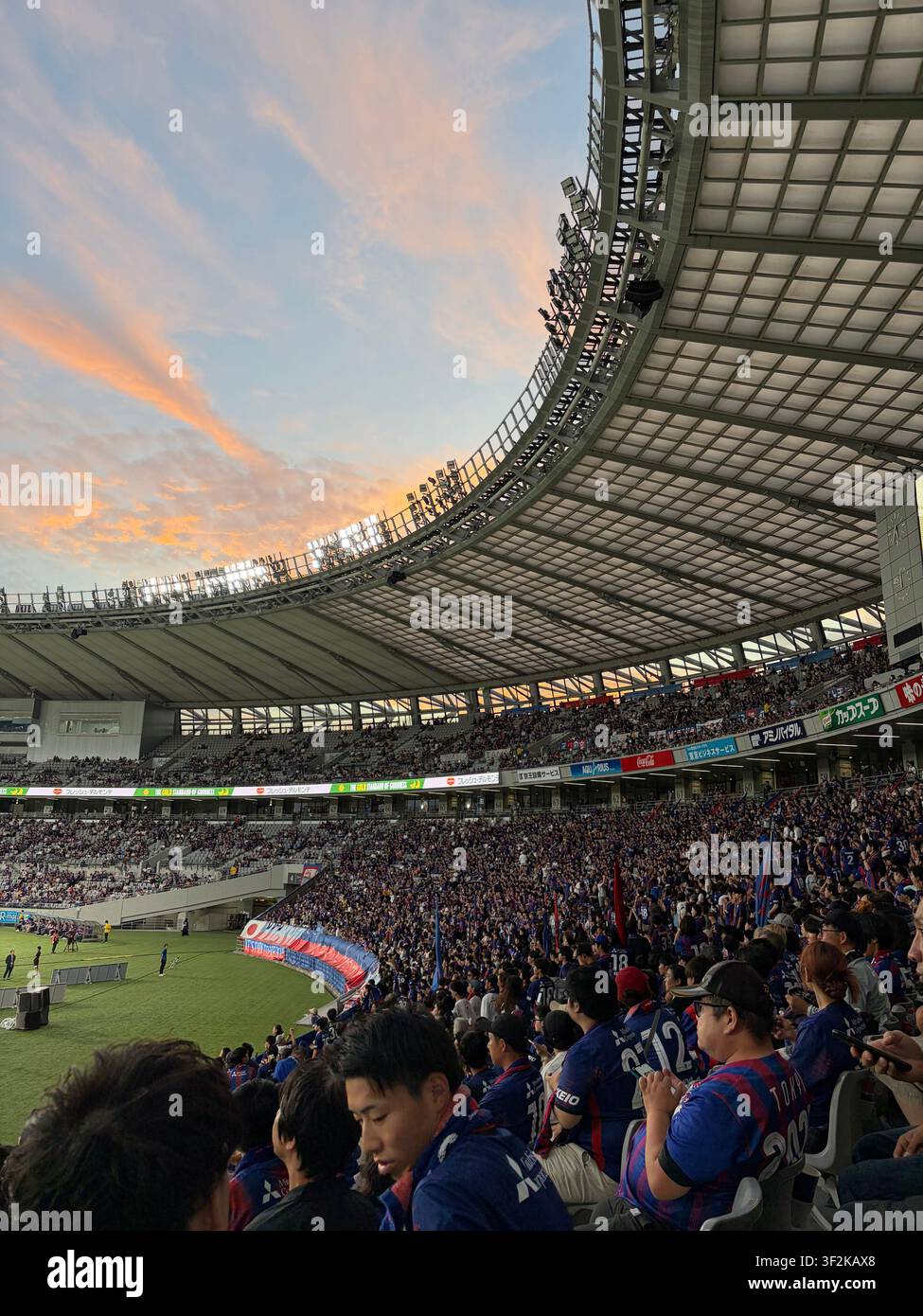 28. September 2025, Inagi, Tokio, Japan – FC-Tokyo-Anhänger füllen die Stadiontribüne bei Sonnenuntergang während eines J-League-Spiels, dramatischer orangefarbener Himmel durch das offene Dach sichtbar. Stockfoto