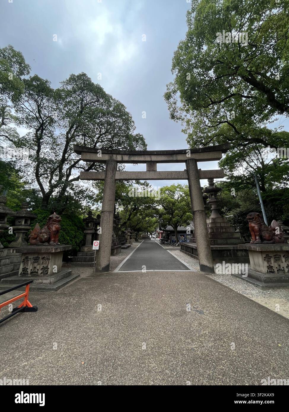 19. Juli 2023, Osaka, Japan – Steinerner Torii-Tor flankiert von Komainu-Wachlöwenhunden an einem traditionellen Shinto-Schrein-Eingang, grüne Bäume und bewölkter Himmel. - Smartphone-aufgenommenes Stockfoto