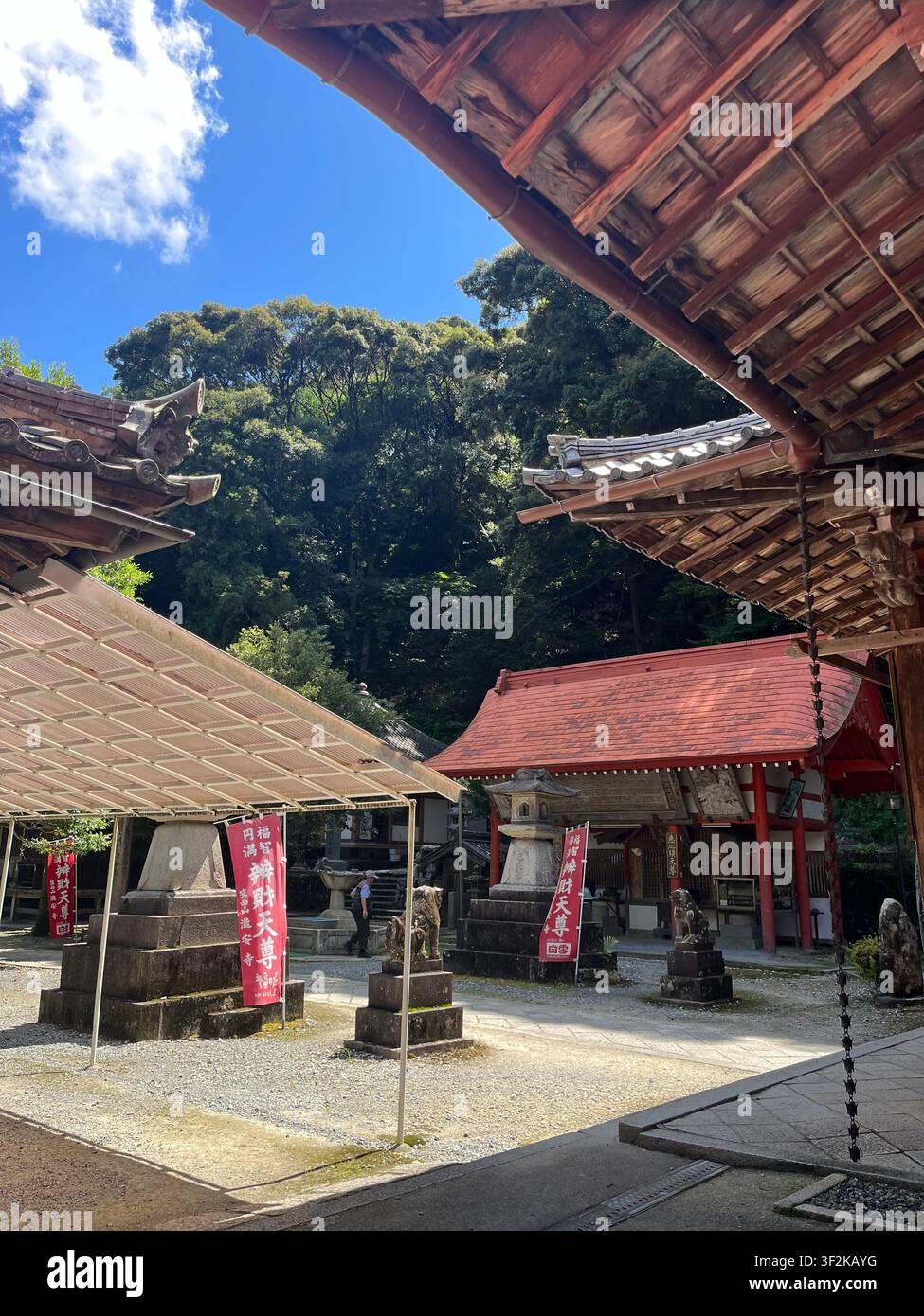 20. Juli 2023, Minoh, Japan – Traditioneller Shinto-Schreinhof mit rotem Torii-Tor, Holzarchitektur, roten Bannern mit japanischem Text und uralten Zedernbäumen. - Smartphone-aufgenommenes Stockfoto