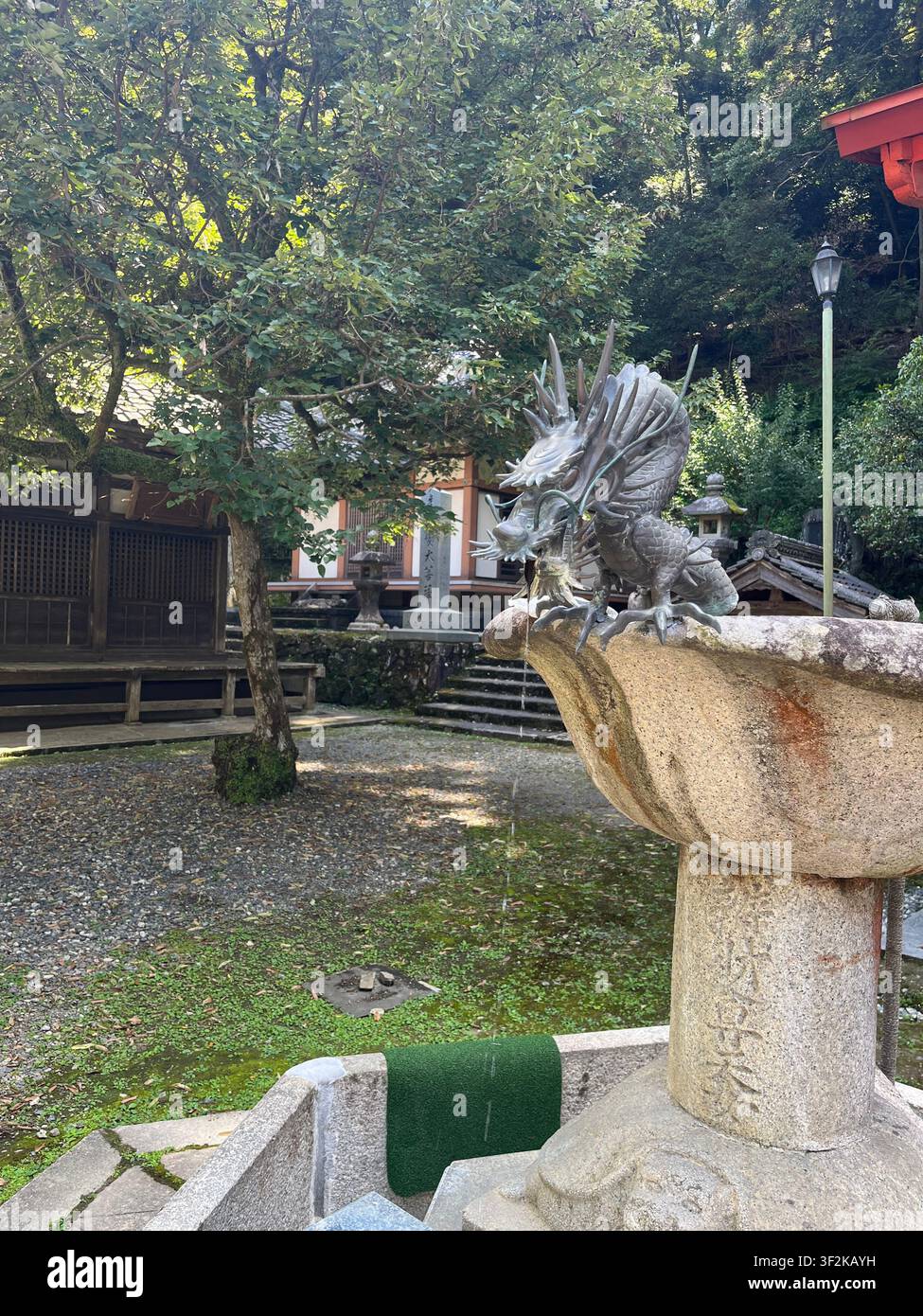 20. Juli 2023, Minoh, Japan – Silberne Drachenstatue und steinerner Laterne mit japanischen Inschriften an einem Shintoschrein in Minoh, Präfektur Osaka, umgeben von üppigen Bäumen. - Smartphone-aufgenommenes Stockfoto