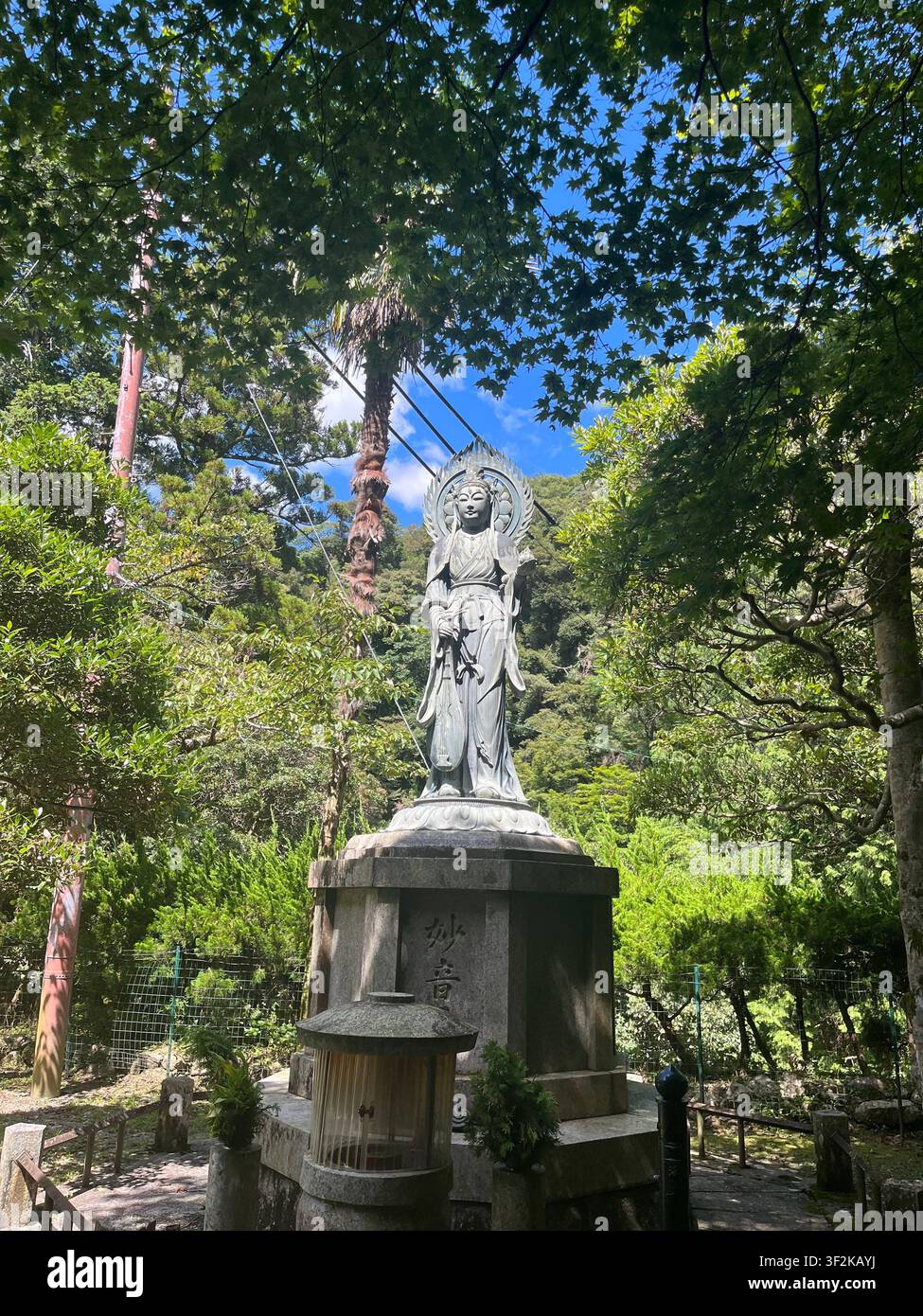 20. Juli 2023, Minoh, Japan – Buddhistische Kannon-Bodhisattva-Steinstatue, umgeben von üppigem grünen Waldkronen und blauem Himmel im Tempel von Minoh, Präfektur Osaka. - Smartphone-aufgenommenes Stockfoto