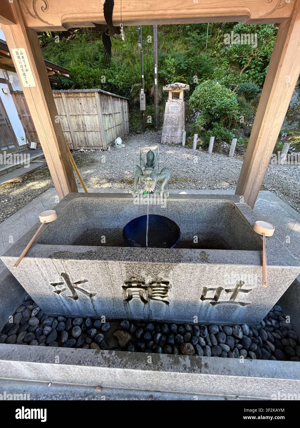 20. Juli 2023, Minoh, Japan – Granit-Temizuya-Ritualwaschbecken mit geschnitzten Kanji-Zeichen und blauer Schale an einem Shinto-Schrein, mit Tengu-Statue und hölzernem Torii-Tor im Hintergrund. - Smartphone-aufgenommenes Stockfoto