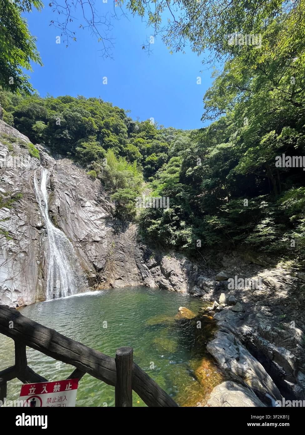 22. Juli 2023, Kobe, Japan – Natürlicher Wasserfall stürzt über moosige Felsen in ein klares Becken, japanisches Warnschild und Holzzaun im Vordergrund, üppiger grüner Wald und blauer Himmel. - Smartphone-aufgenommenes Stockfoto