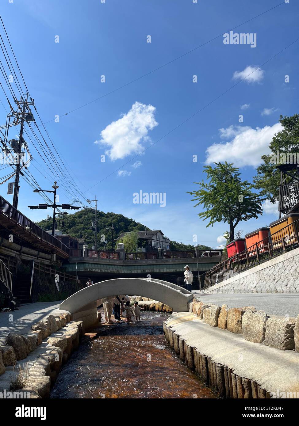 23. Juli 2023, Kobe, Japan – Traditionelle Straßenansicht des Arima-Onsen-Badespringsviertels mit Steinbrücke, japanischen Schildern, Stromleitungen und üppigen Sommerbäumen. - Smartphone-aufgenommenes Stockfoto