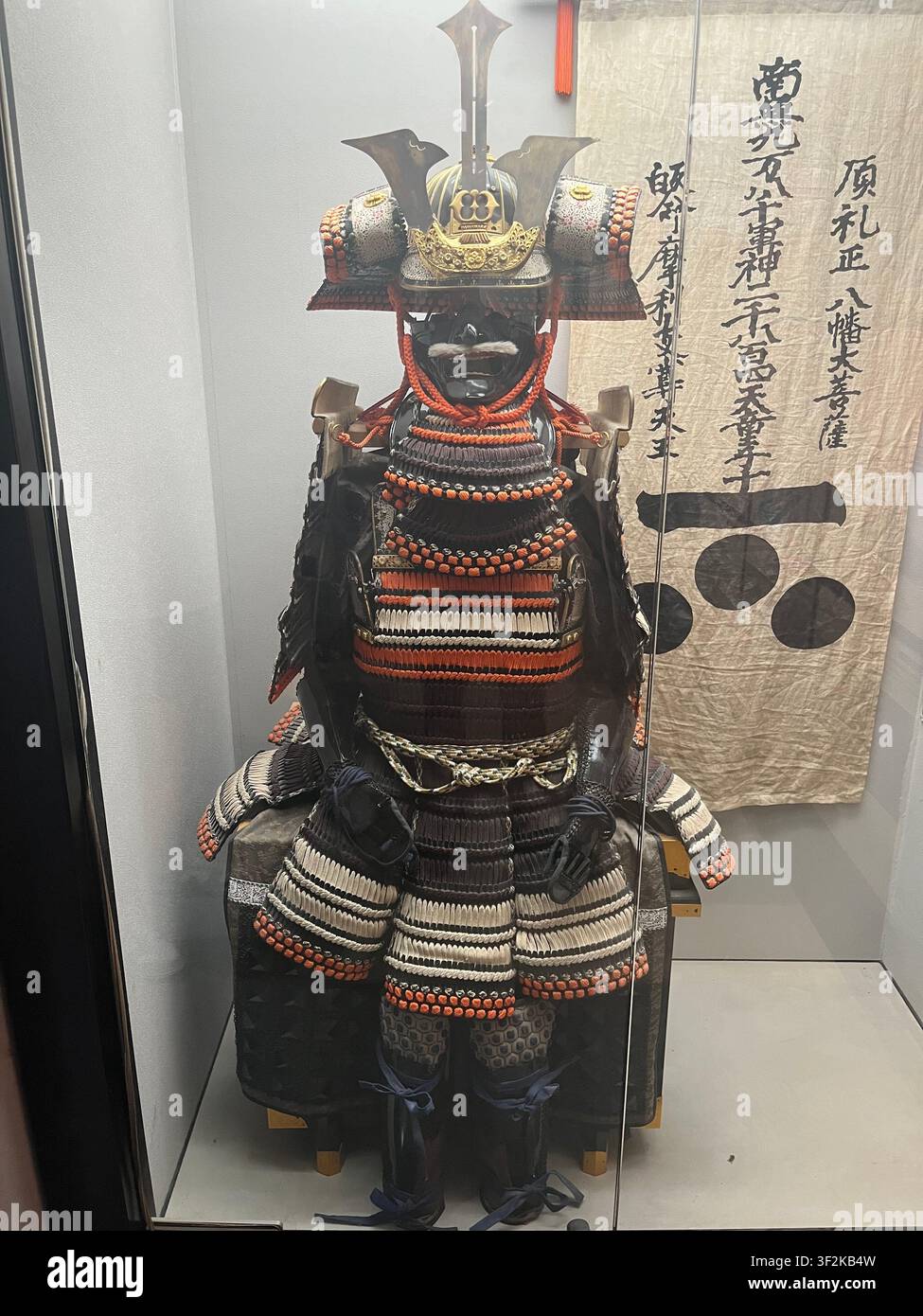 25. Juli 2023, Hiroshima, Japan – Vollständige traditionelle Samurai-Rüstung (yoroi) in einer Museumsvitrine ausgestellt mit kalligraphischem japanischem Schriftrollenbanner im Hintergrund. - Smartphone-aufgenommenes Stockfoto