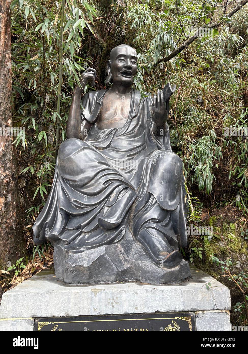 23. März 2025, Bán Bó, Provinz Lai Châu, Vietnam - Dunkle bronzene liegende Buddha-Statue, umgeben von wilder Bergvegetation mit vietnamesischer Gedenktafel. - Smartphone-aufgenommenes Stockfoto