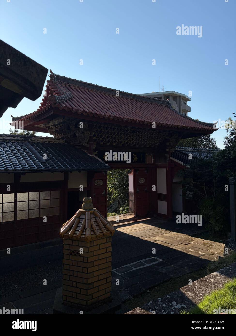 11. Oktober 2025, Nagasaki, Japan – Rot lackiertes Tempeltor mit Kanji-Inschrift und kunstvoll geschwungenem Ziegeldach, Blick von unten gegen klaren blauen Himmel. - Smartphone-aufgenommenes Stockfoto