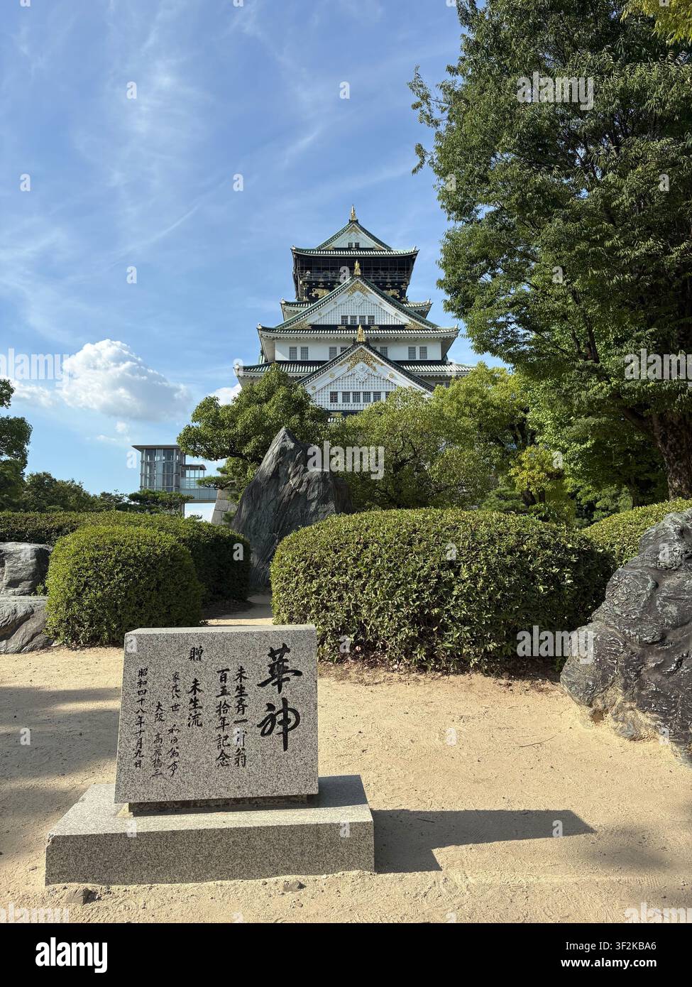 28. Juli 2025, Osaka, Japan – Der Donjon von Osaka-Schloss erhebt sich hinter gepflegten Hecken und einem geschnitzten japanischen Steinmonument unter einem sommerlichen blauen Himmel. - Smartphone-aufgenommenes Stockfoto