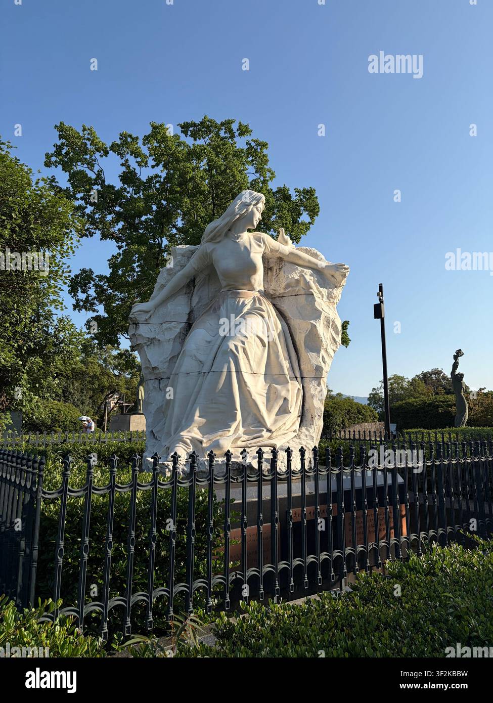 12. Oktober 2025, Nagasaki, Japan – Weiße Marmorreligionsstatue mit fließenden Gewändern vor klarem blauem Himmel, kunstvollem Eisengitter, an einem historischen katholischen Ort in Nagasaki. - Smartphone-aufgenommenes Stockfoto