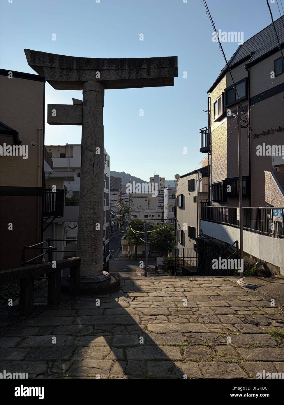 12. Oktober 2025, Nagasaki, Japan – Steinerner Torii-Tor eines städtischen Shinto-Schreins, von einem niedrigen Winkel aus fotografiert, mit Stadtgebäuden und klarem Himmel im Hintergrund. - Smartphone-aufgenommenes Stockfoto