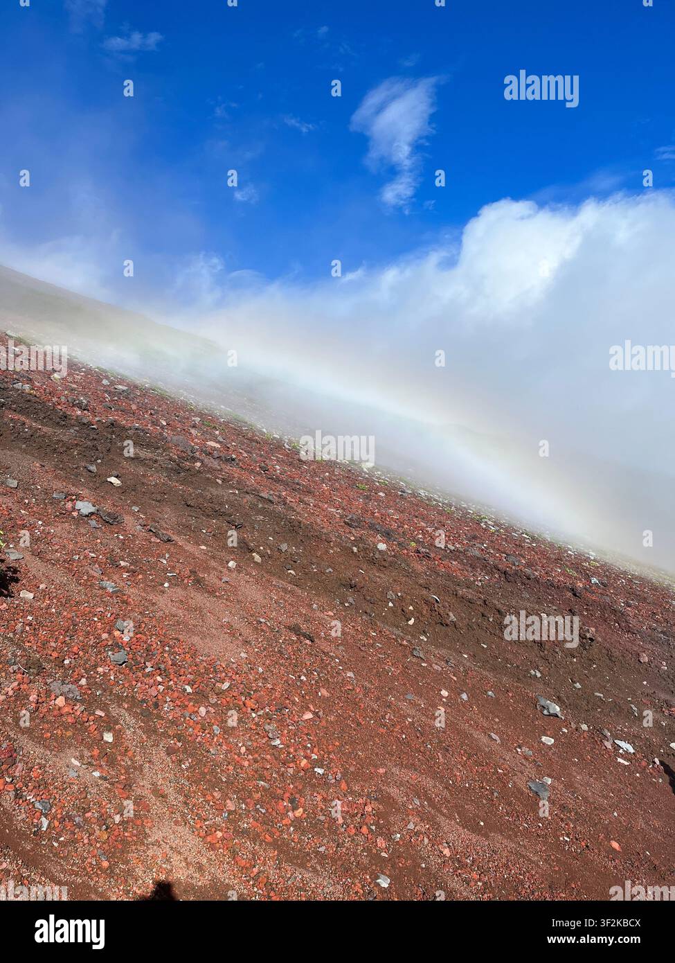 Roter vulkanischer Kies und Felshang des Berges Fuji (Oyama) mit dramatischem Regenbogen und Wolkennebel vor einem lebendigen blauen Himmel, Shizuoka, Japan. - Smartphone-aufgenommenes Stockfoto