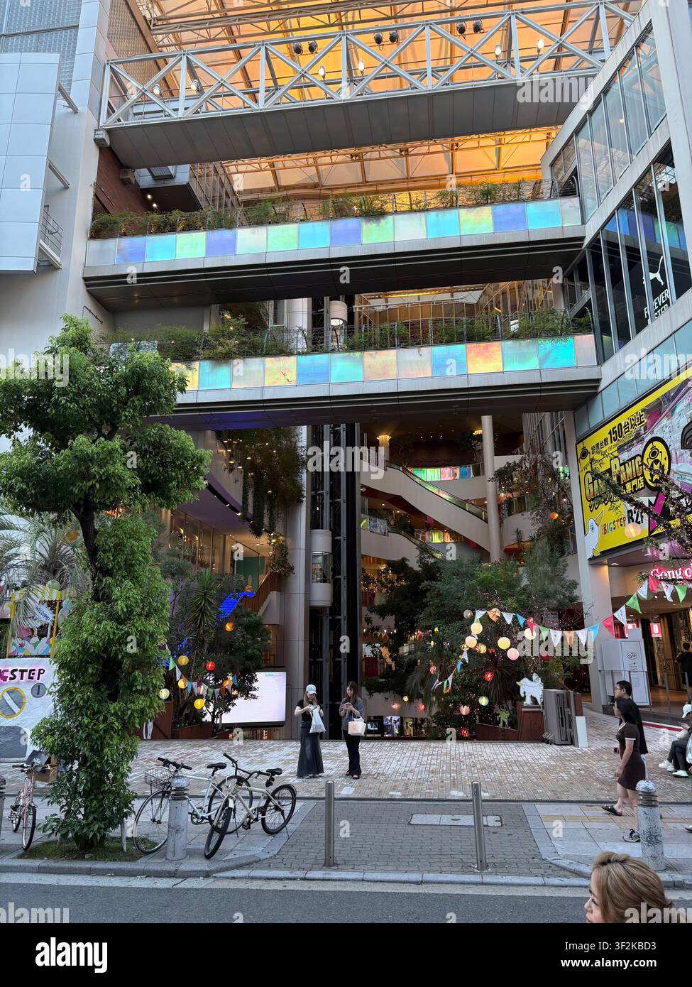 28. Juli 2025, Osaka, Japan – Moderner mehrstöckiger Einkaufskomplex mit irisierenden Glasflächen, grünen Bäumen, bunten Laternen und Fußgängern im Stadtzentrum von Osaka. - Smartphone-aufgenommenes Stockfoto