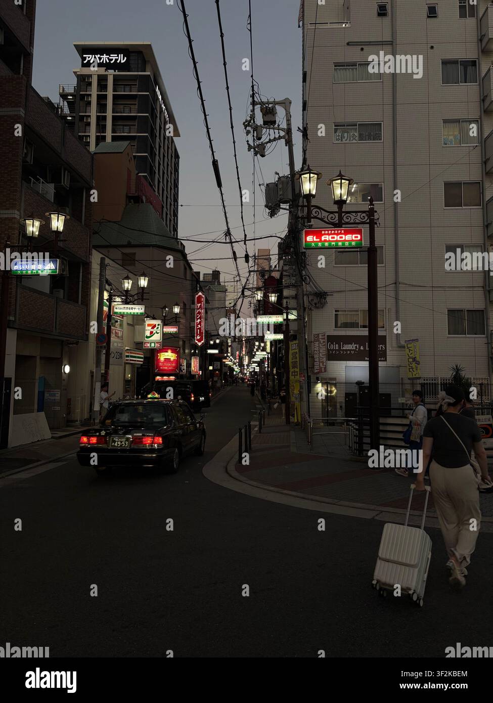 28. Juli 2025, Osaka, Japan – Stadtstraßenszene in der Dämmerung mit leuchtenden Neonschildern, japanischen Ladengeschäften, Stromleitungen und Fußgängern in der Innenstadt von Osaka. - Smartphone-aufgenommenes Stockfoto