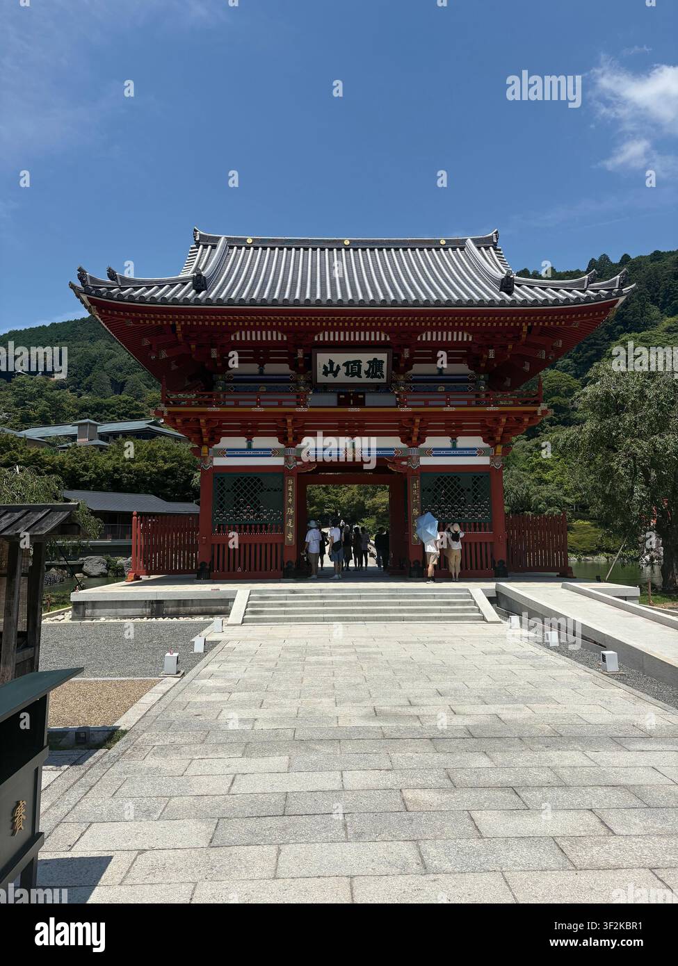 30. Juli 2025, Minoh, Japan – Traditionelles rotes buddhistisches Tempeltor (sanmon) mit japanischer Kanji-Inschrift, Besucher gehen hindurch, üppige grüne bewaldete Hügel im Hintergrund. - Smartphone-aufgenommenes Stockfoto