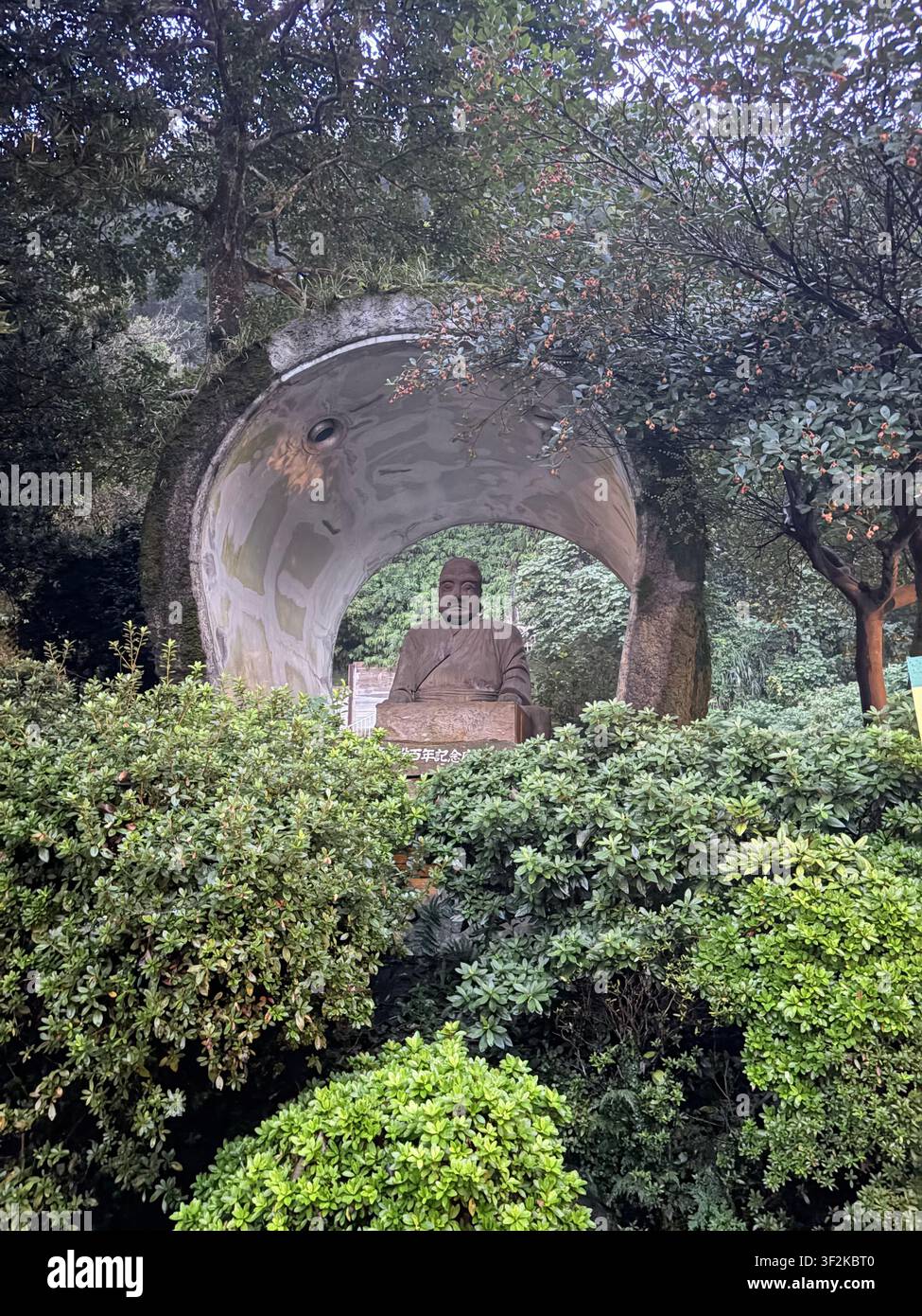 19. Oktober 2025, Kagoshima, Japan – Bronzebüste auf einem halbmondförmigen Steinpodest, umgeben von dichtem grünem Gartenlaub, mit sichtbarer japanischer Texttafel. - Smartphone-aufgenommenes Stockfoto