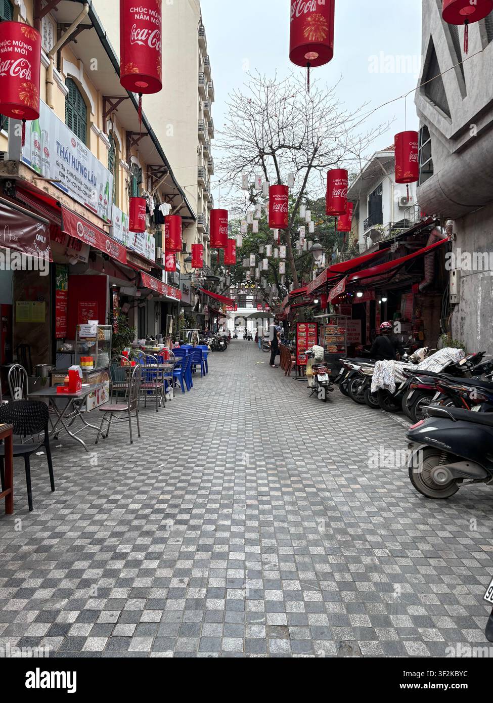 26. März 2025, Hanoi, Vietnam – Straßenrestaurant, das mit Coca-Cola-Tet-Laternen und vietnamesischer Neujahrsmarken entlang eines belebten Gehwegs in Hanoi geschmückt ist, mit Motorrädern. - Smartphone-aufgenommenes Stockfoto