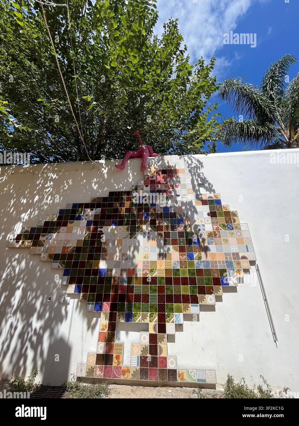 7. September 2023, Mittelmeerregion - Außenwandinstallation mit einem großen Pixel-Art-Fliesenmosaik mit rosa Kletterfiguren-Skulptur, gesprenkelten Baumschatten, blauem Himmel. - Smartphone-aufgenommenes Stockfoto