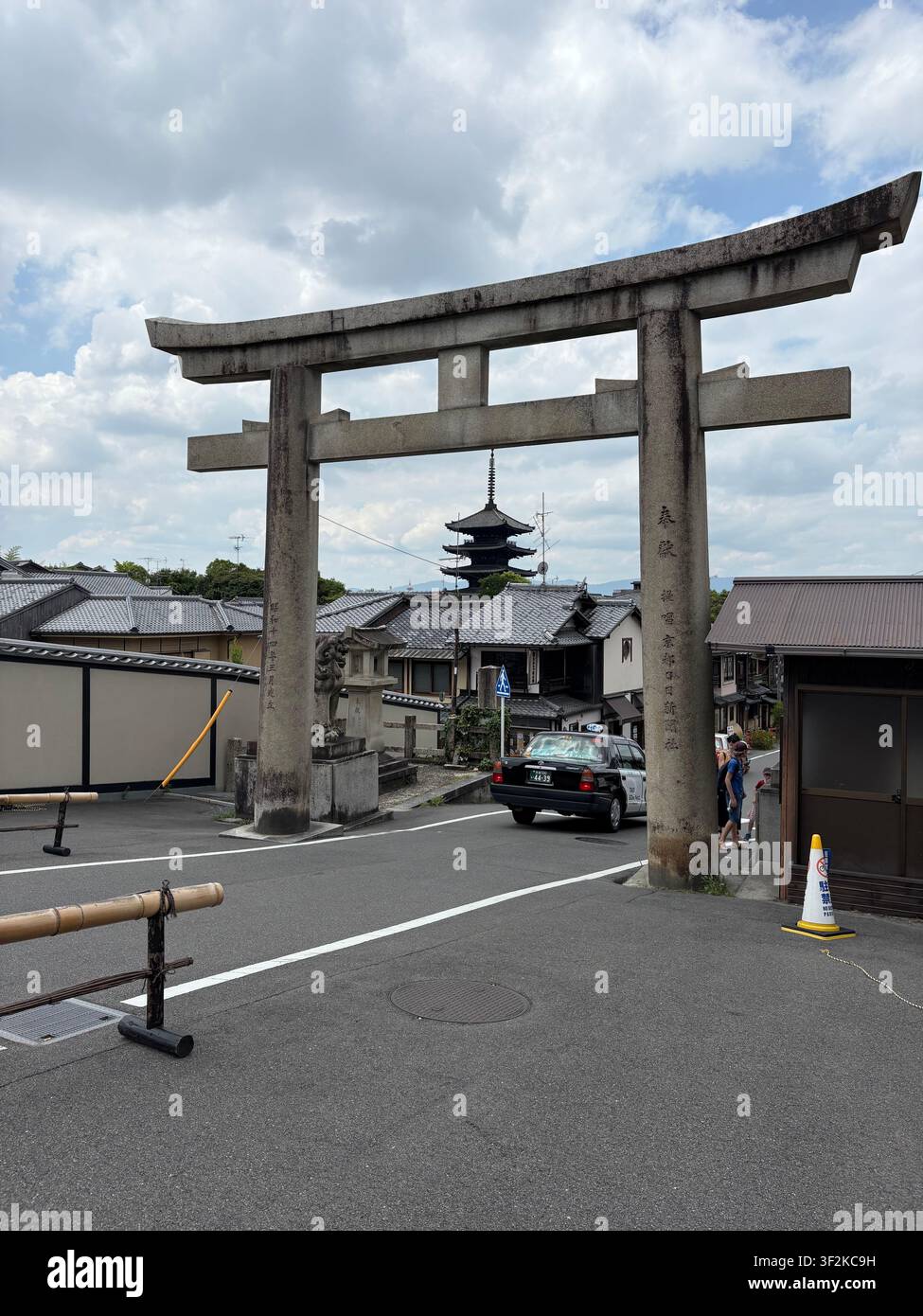 3. August 2025, Kyoto, Japan – Steinerner Torii-Torbogen mit japanischen Kanji-Inschriften, der eine traditionelle Pagode und eine urbane Straßenszene im sommerlichen Kyoto einrahmt. - Smartphone-aufgenommenes Stockfoto