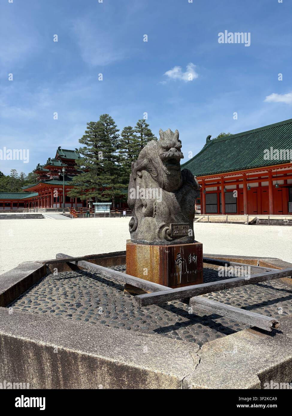 4. August 2025, Kyoto, Japan – Steatue eines steinernen Löwenhundes Komainu auf einem Sockel an einem traditionellen japanischen Shintō-Schrein mit roter Pagodenarchitektur im Hintergrund. - Smartphone-aufgenommenes Stockfoto