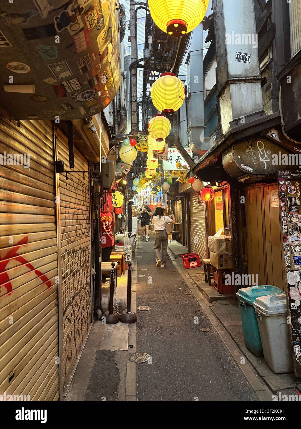 13. August 2025, Shibuya, Japan – Schmale Gasse unter Bahngleisen, gesäumt von leuchtenden Chōchin-Laternen und Izakaya-Restaurants, Sonnenblumen und Aufkleber verleihen urbaner Struktur. - Smartphone-aufgenommenes Stockfoto