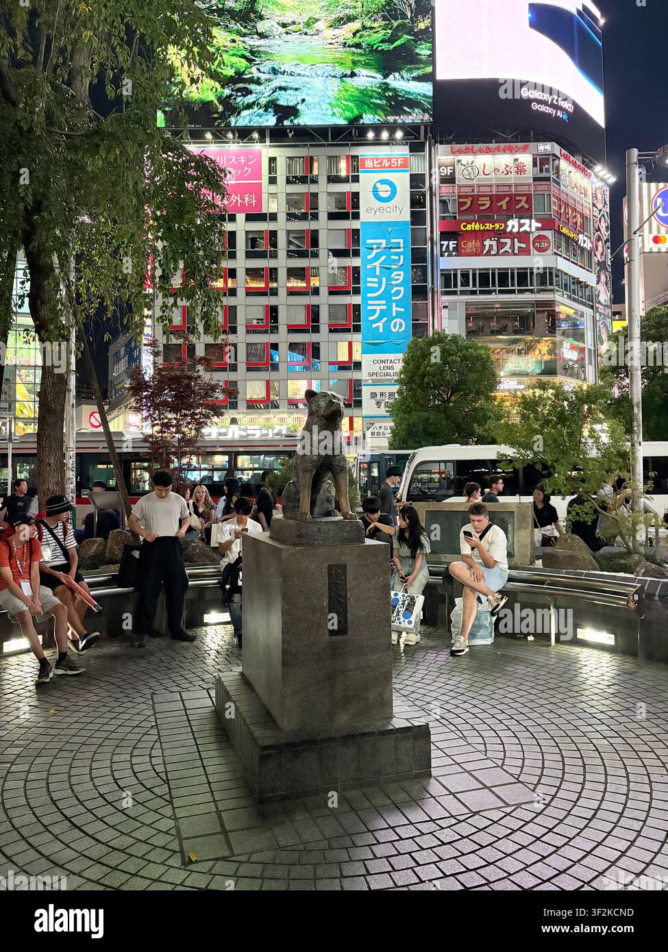 13. August 2025, Shibuya, Tokio, Japan – Touristen versammeln sich nachts um die ikonische Hachiko-Hundestatue vor dem Shibuya-Bahnhof, umgeben von hellen japanischen Neonreklamen. - Smartphone-aufgenommenes Stockfoto
