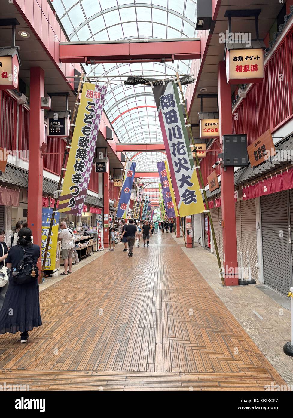 14. August 2025, Japan – Überdachte Einkaufsstraße mit großen bunten japanischen Festbannern, die von der Decke hängen, während Käufer unter traditioneller roter Architektur vorbeigehen. - Smartphone-aufgenommenes Stockfoto