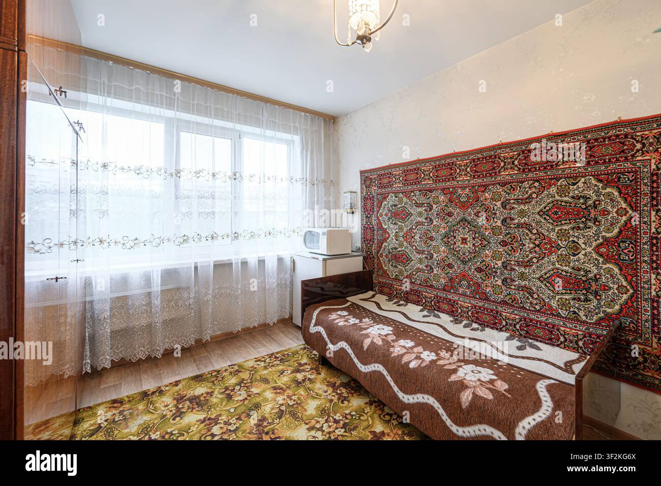 Gemütliches Zimmer mit einer Couch an einer teppichbedeckten Wand. Ein Fenster mit durchsichtigen Vorhängen lässt Licht herein. Ein kleiner Kühlschrank und eine Mikrowelle stehen neben der Couch Stockfoto