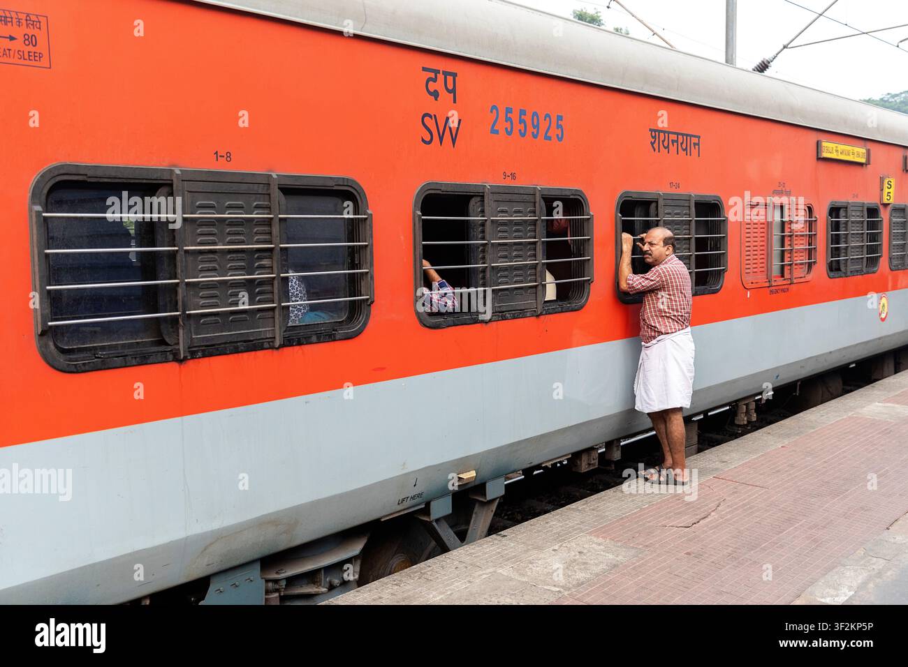 Mann hält sich an den Schienen des Fensters eines orangefarbenen Zugwagens auf einem Bahnsteig während der Fahrt in Kerala, Indien. Stockfoto