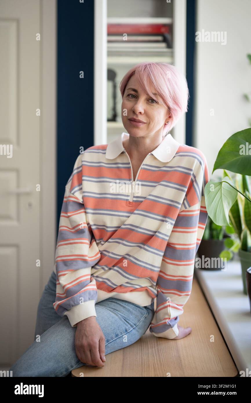 Entspannte, positive, stilvolle reife Frau, die vor der Kamera posiert, während sie auf einem Tisch neben dem Fenster zu Hause sitzt. Stockfoto