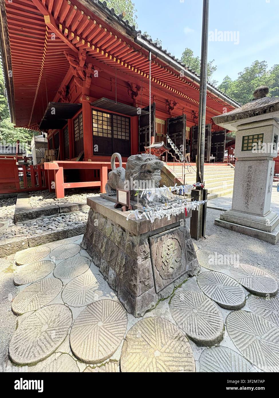 17. August 2025, Nikko, Japan – Steinerne Komainu-Wächterstatue mit Gebetsgaben neben einem roten Holzschrein und einem geschnitzten Steinlaternen am UNESCO-Weltkulturerbestandort Nikko. - Smartphone-aufgenommenes Stockfoto