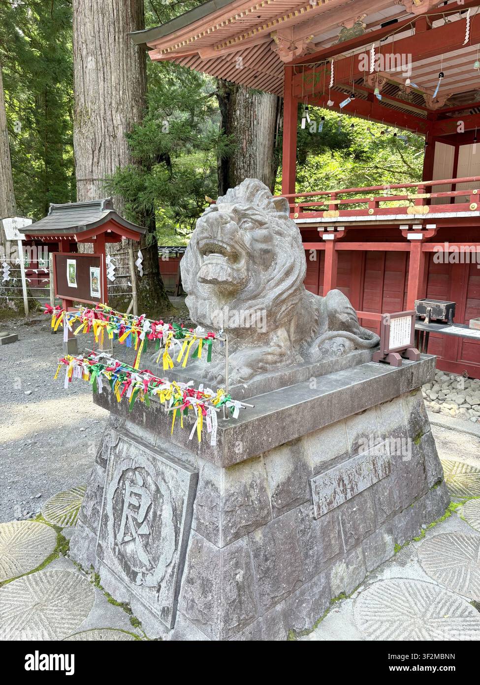 17. August 2025, Nikko, Japan – Steinerne Komainu-Wachlöwenstatue, geschmückt mit bunten Tanzaku-Papiergaben, an einem UNESCO-Shintoschrein in Nikko. - Smartphone-aufgenommenes Stockfoto