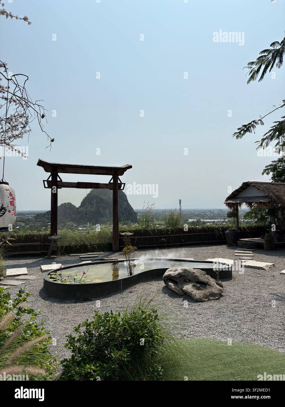 7. April 2025, Da Nang, Vietnam – Traditioneller asiatischer Garten mit ovalem Lotus-Teich, dekorativem Felsen, hölzerner Torstruktur und nebligen Kalksteinbergen in der Dämmerung. - Smartphone-aufgenommenes Stockfoto