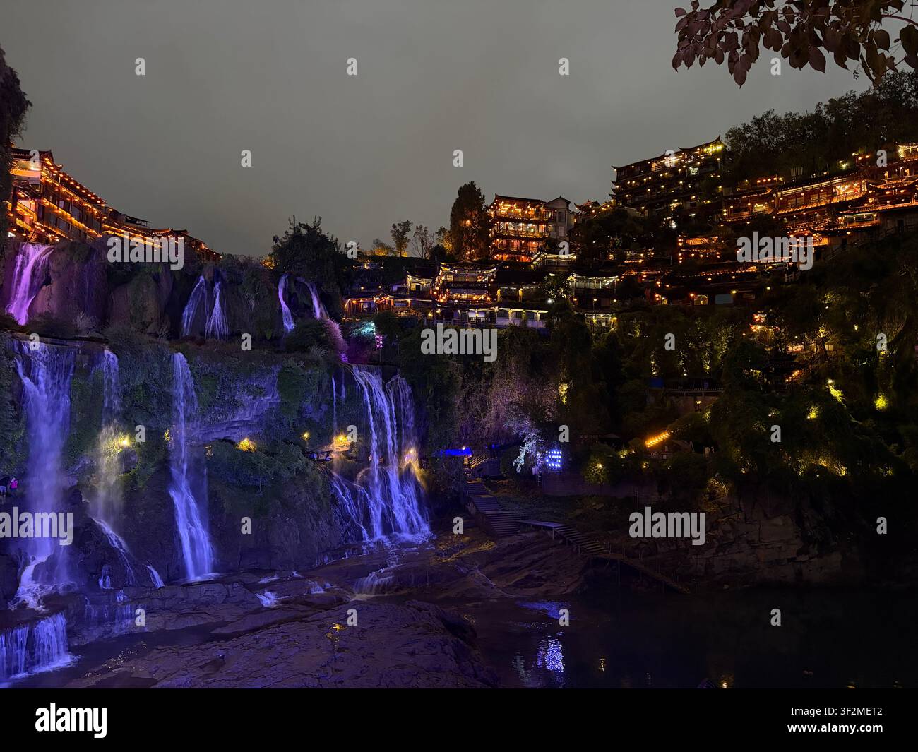 Beleuchtete Kaskaden-Wasserfälle und traditionelle chinesische Pfahlhäuser bei Nacht in Furong Town (Furong Zhen), Guzhang, Xiangxi, Hunan-Provinz, China. - Smartphone-aufgenommenes Stockfoto