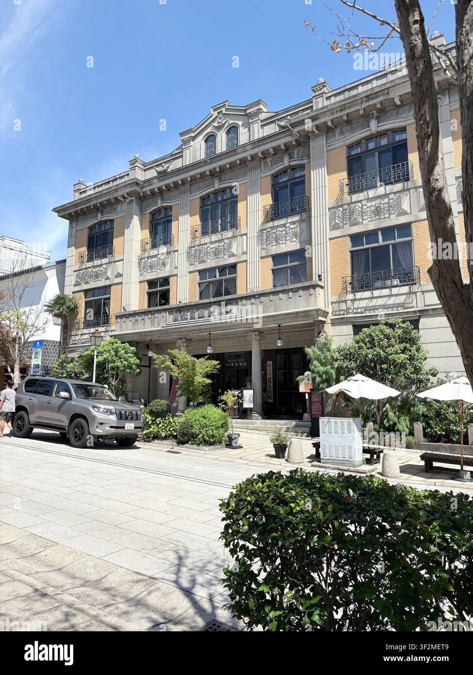 28. August 2025, Nagano, Japan – Historisches Gebäude im westlichen Stil aus der Meiji-Zeit an einer sonnigen Straße in Nagano mit kunstvoller Fassade, japanischer Beschriftung und geparktem SUV. - Smartphone-aufgenommenes Stockfoto