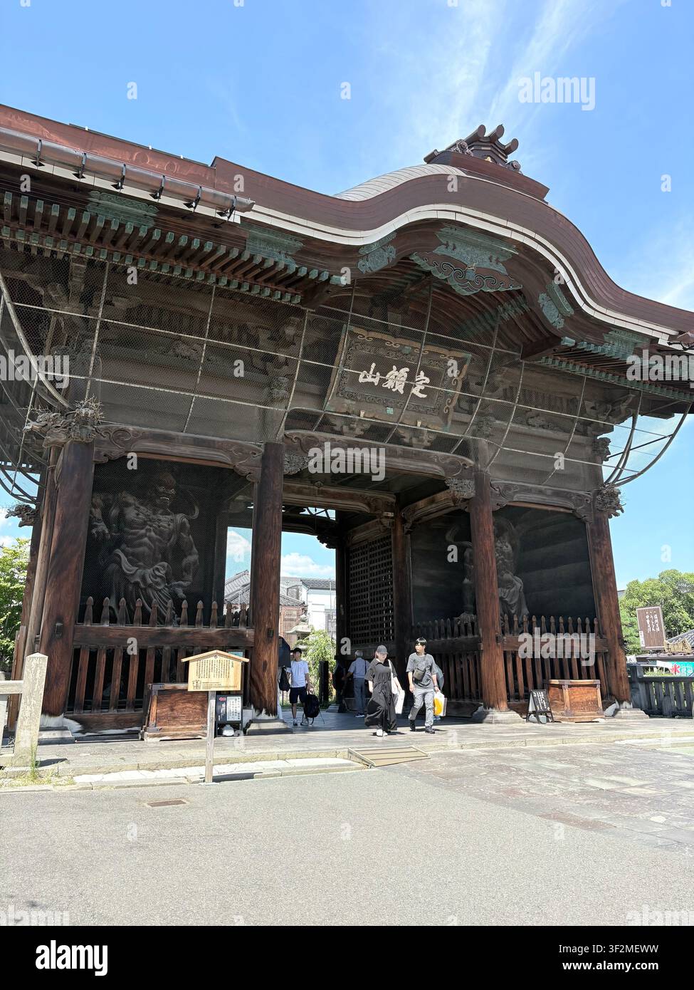 28. August 2025, Nagano, Japan – Verschnörkeltes hölzernes Sanmon-Tor des Zenkoji-Buddhistentempels mit Kanji-Plakette, Besuchern und dekorativen Metallmaschen-Details unter blauem Himmel. - Smartphone-aufgenommenes Stockfoto