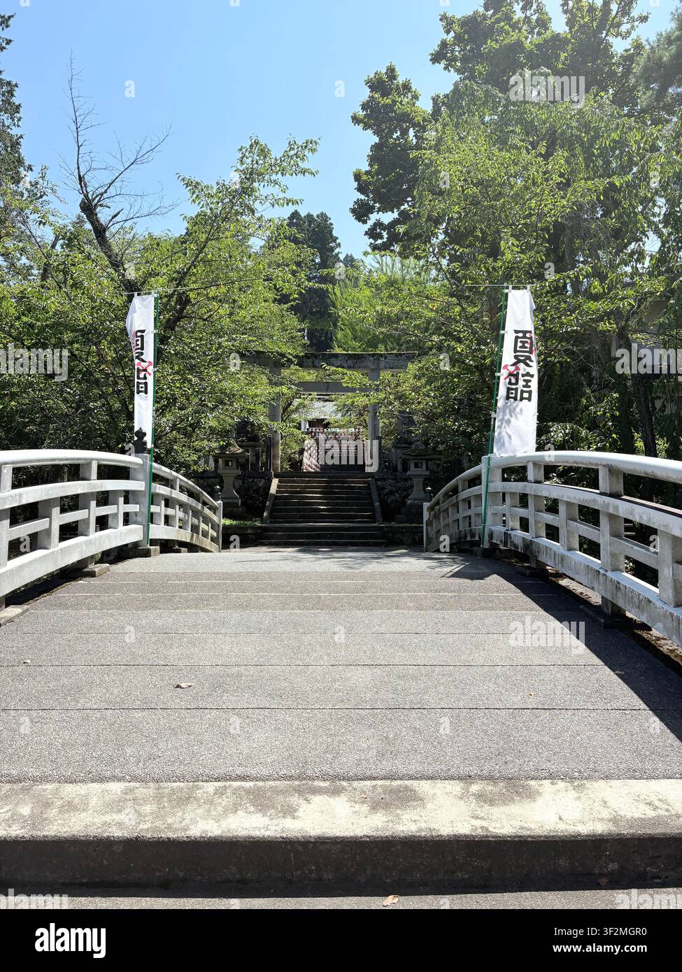 31. August 2025, Takayama, Japan – Steinerner Torii-Tor mit japanischen Kanji-Bannern am Eingang eines Shintō-Schreins, umgeben von grünen Bäumen im Sommer. - Smartphone-aufgenommenes Stockfoto